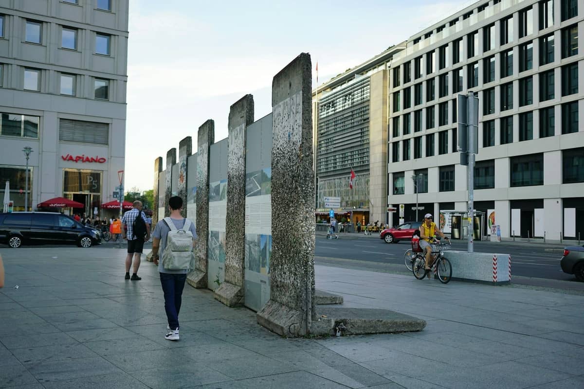 Segmente der Berliner Mauer mit historischen Fotos darauf, aufgestellt in einer städtischen Straßenlandschaft mit Fußgängern in der Nähe.