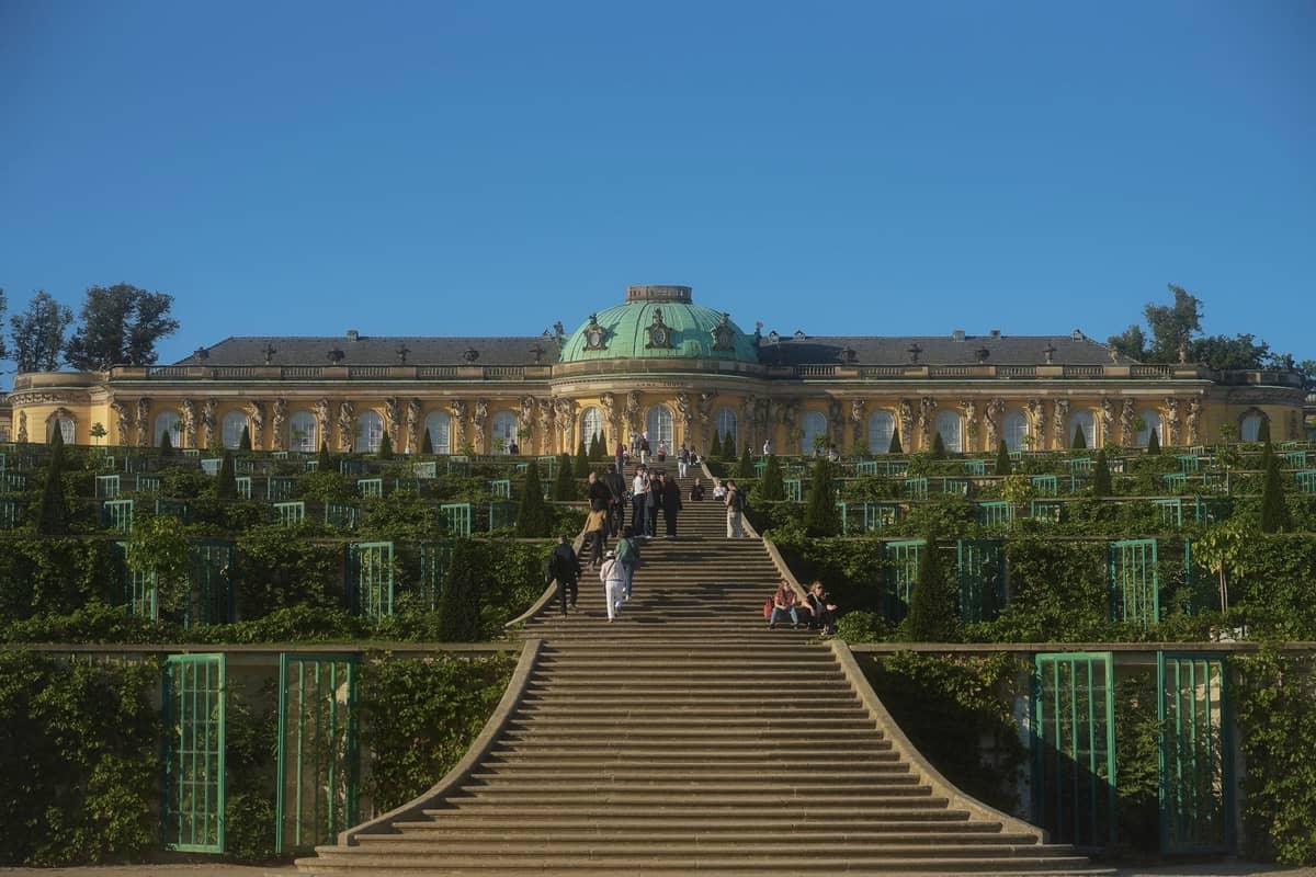 Breite Steintreppe und terrassierte Gärten, die zum prunkvollen Schloss Sanssouci mit grüner Kuppel unter einem klaren blauen Himmel führen.