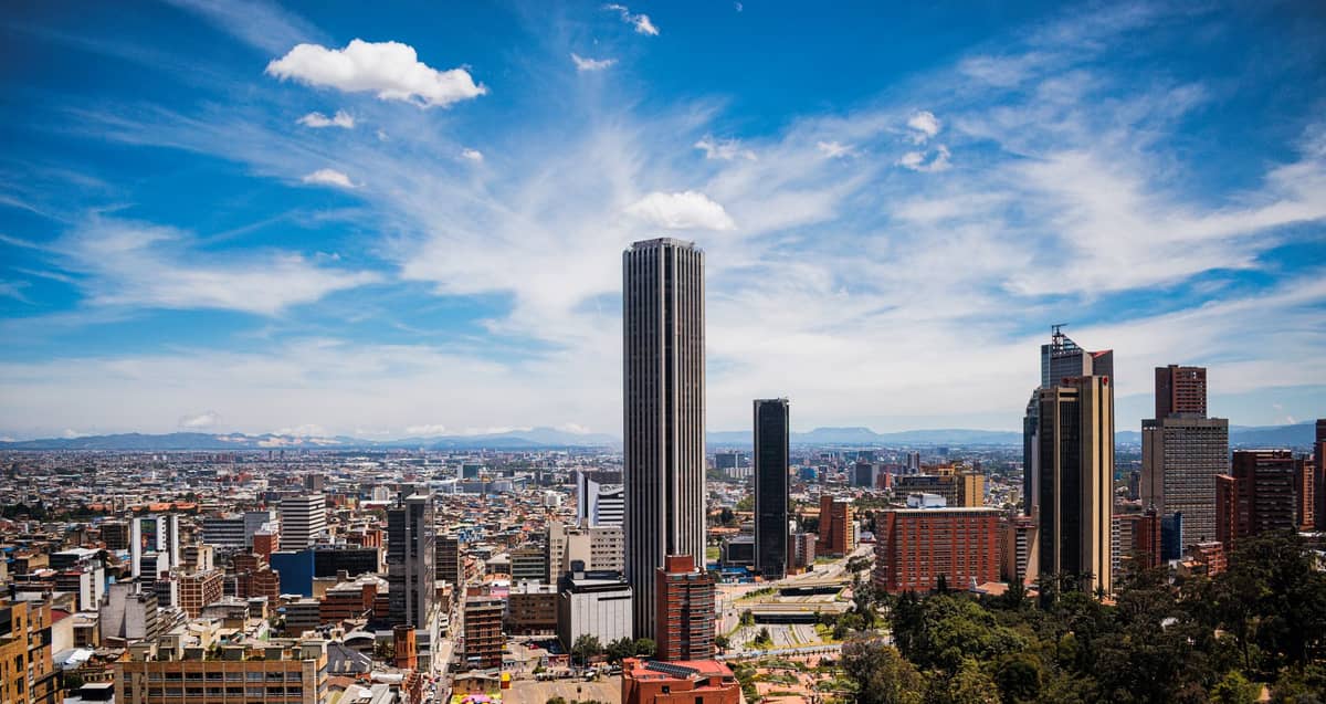Panoramic view of Bogotá city center with tall skyscrapers, mountainous backdrop, and a wide expanse of urban landscape under a dramatic blue sky.
