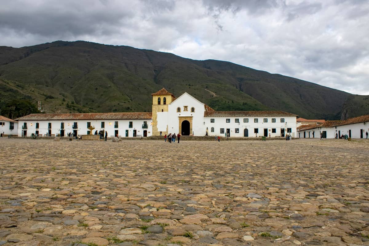 Wide view of the large cobblestone Plaza Mayor of Villa de Leyva with the iconic white church and surrounding colonial buildings, mountains in background.
