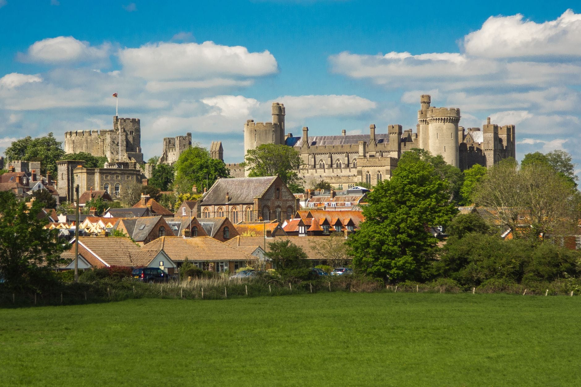 Panoramablick auf eine historische Burg, die über einem Dorf mit roten Dächern aufragt, umgeben von grünen Feldern und Bäumen unter einem blauen Himmel mit Wolken.