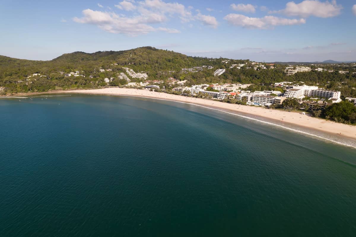 Luftaufnahme von Noosa Main Beach mit klarem blauem Wasser, Sandstrand sowie Hotels und bewaldeten Hügeln im Hintergrund.