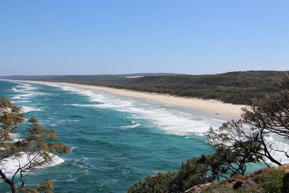 Weitblick auf den Main Beach auf North Stradbroke Island mit türkisfarbenen Wellen, sandiger Küstenlinie und bewaldeten Dünen unter einem klaren blauen Himmel.