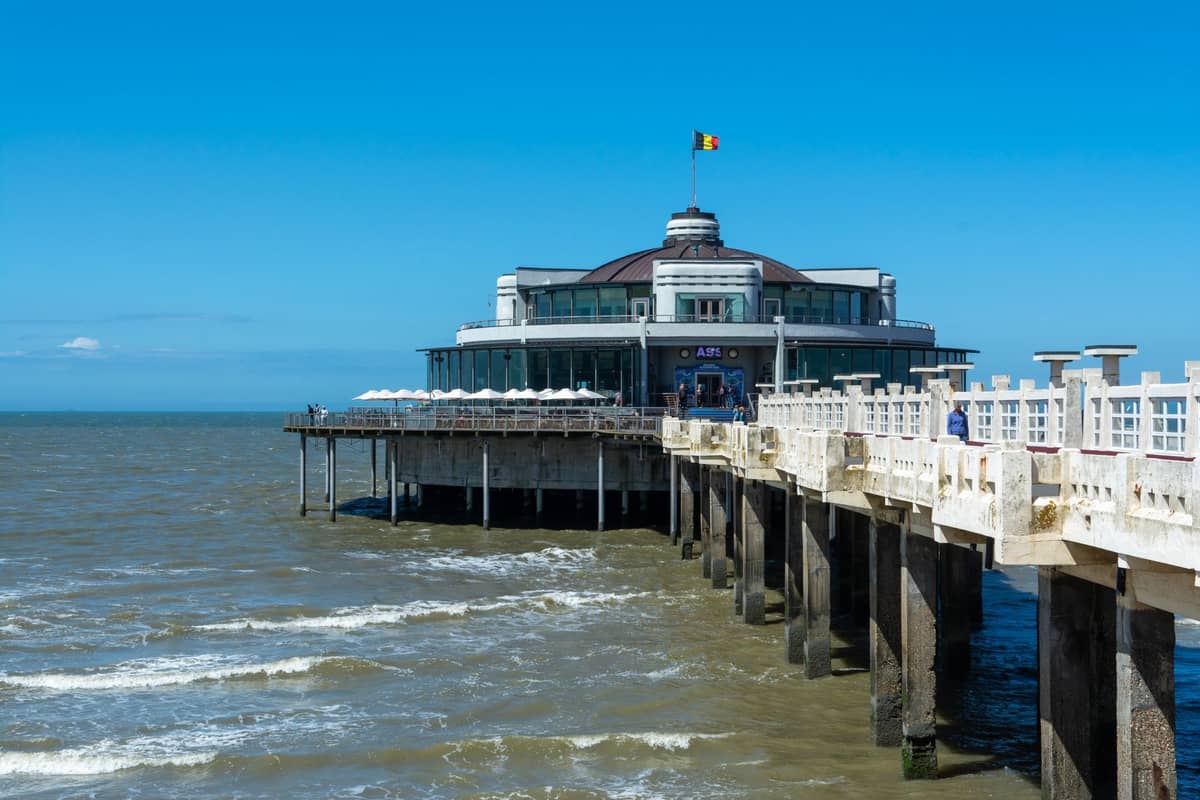 Blankenberge Pier with a Belgian flag flying above the round pavilion at the end, extending into the North Sea under blue skies.