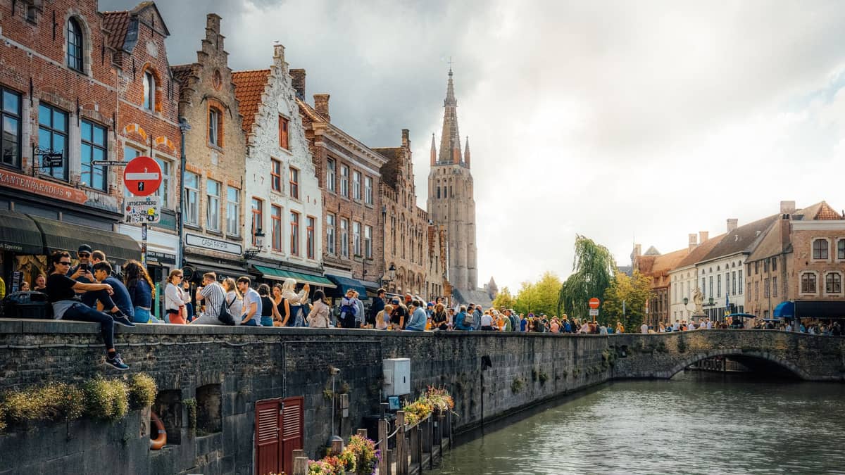 View of Bruges canal lined with historic buildings and Belfry of Bruges, crowds of people along the waterfront under a partly cloudy sky.