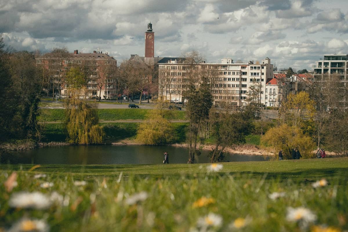 Vista de um parque gramado nas proximidades de Bruxelas com um lago, edifícios residenciais ao fundo e uma torre com relógio sob um céu nublado.