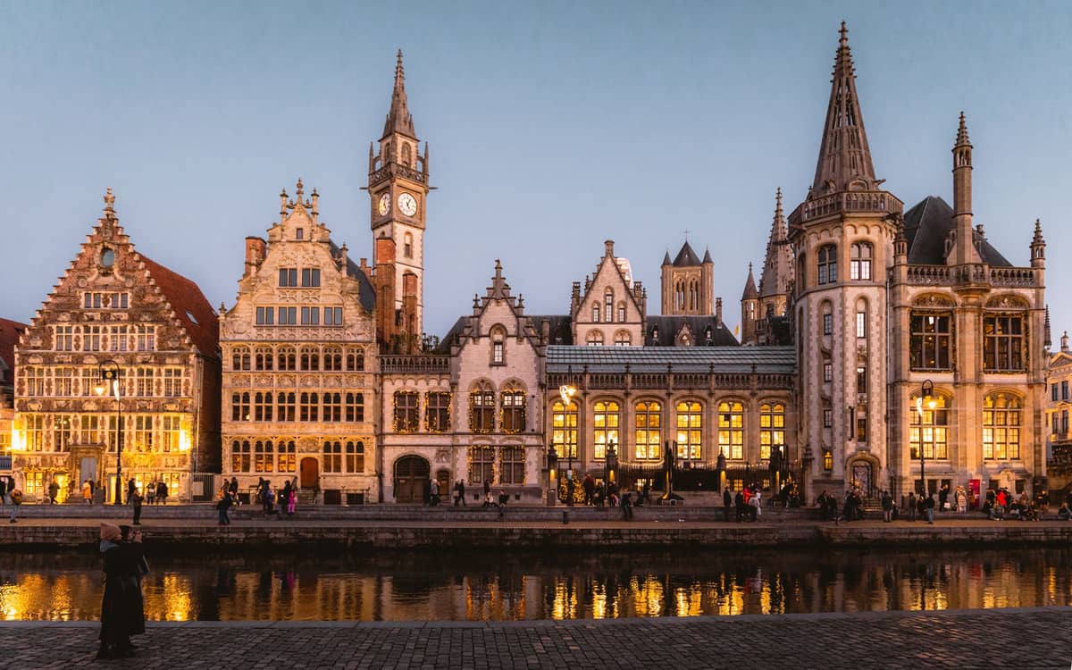 Vista noturna das corporações medievais e da torre do relógio de Ghent iluminadas de forma acolhedora ao longo do canal Graslei com pessoas passeando à beira da água.