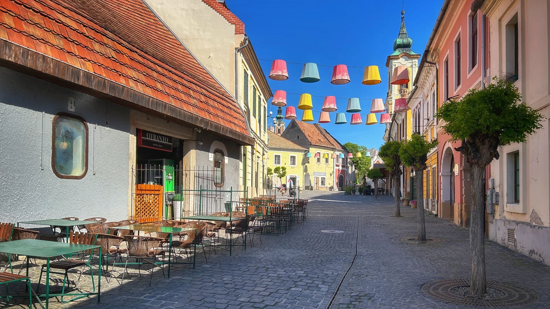 Kolorowe abażury zwisające nad brukowaną ulicą otoczoną pastelowymi budynkami i kawiarniami na świeżym powietrzu w Szentendre w słoneczny dzień.