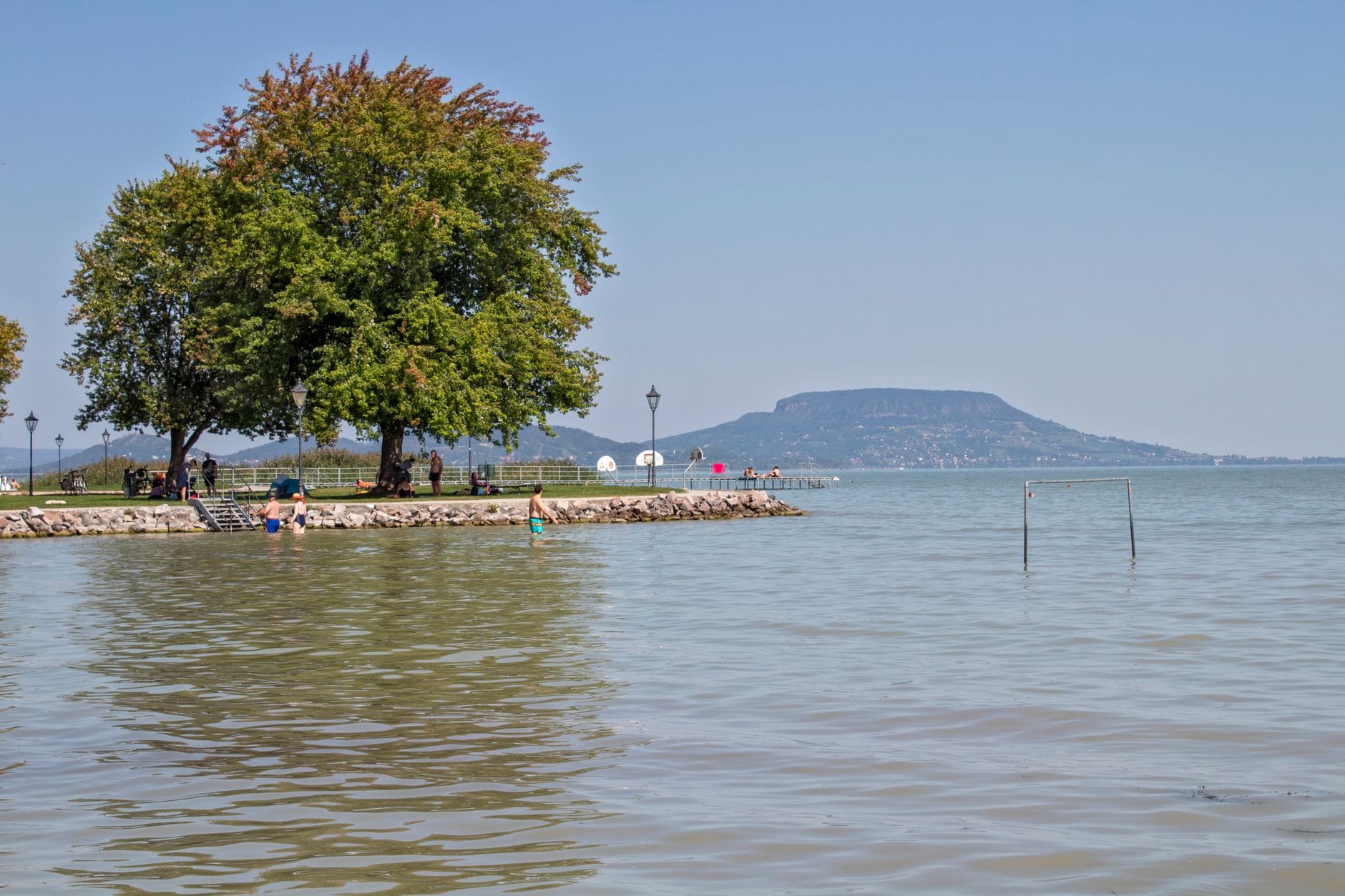 Jezioro Balaton z zalesioną promenadą, spokojną wodą, ludźmi wypoczywającymi na brzegu i charakterystycznym wzgórzem Badacsony w tle.