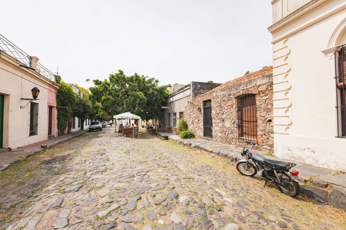 ถนนปูหินในย่านประวัติศาสตร์อาณานิคมของ Colonia del Sacramento เรียงรายด้วยอาคารหินและปูนเก่าแก่พร้อมรถมอเตอร์ไซค์จอดอยู่