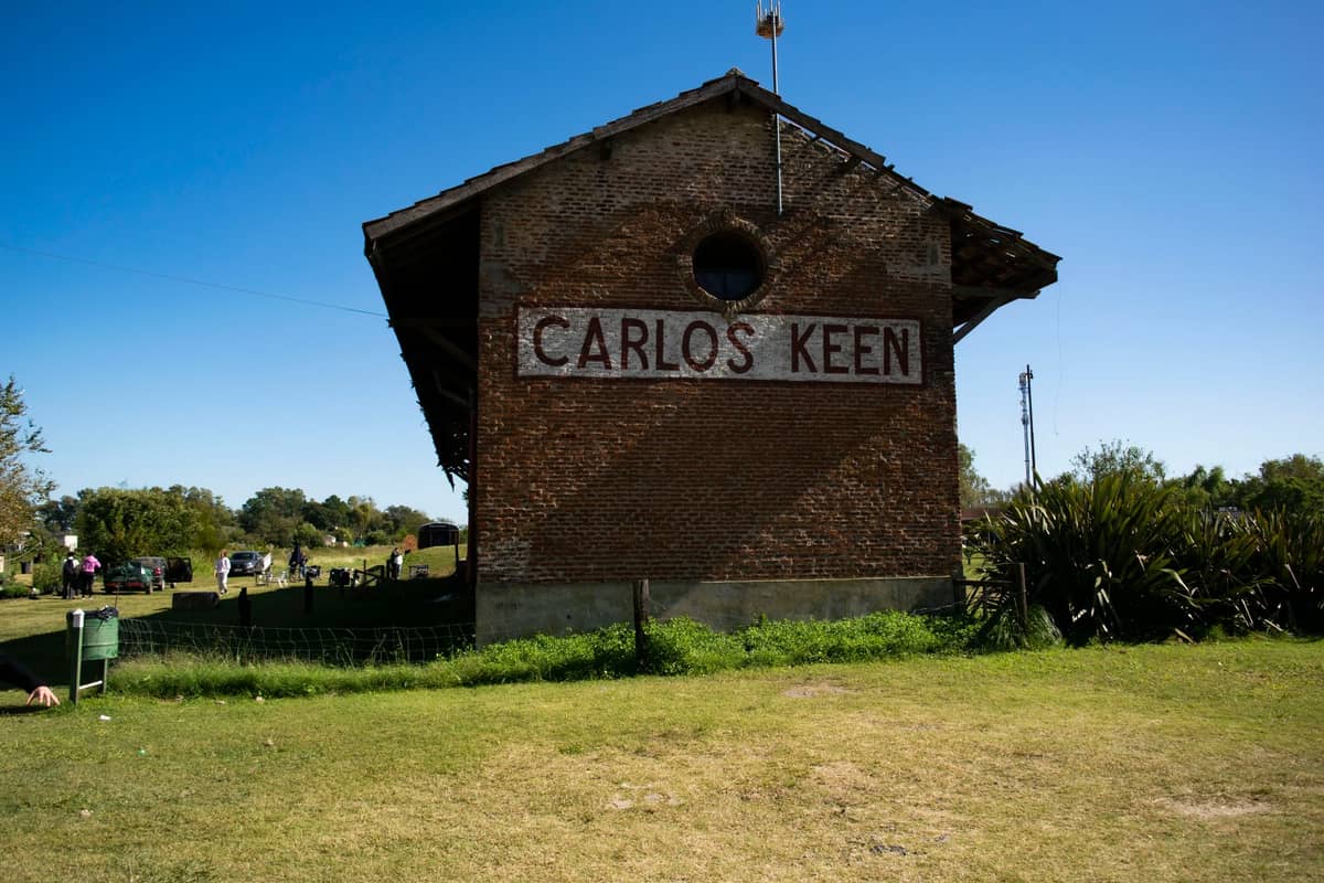 สถานีรถไฟอิฐเก่าที่ Carlos Keen พร้อมป้ายหน้าตึก สนามหญ้า และท้องฟ้าสีฟ้า หนึ่งในทริปวันเดียวสุดฮิตจากบัวโนสไอเรส
