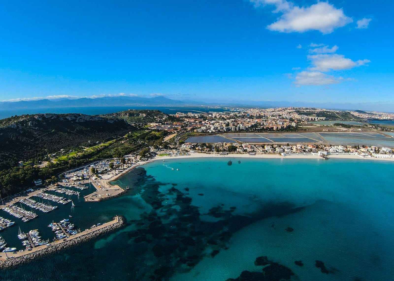 Luftaufnahme von Cagliari mit türkisfarbenem Wasser, Marina, Küstenlinie und Stadtpanorama unter strahlendem Himmel – ein Blick auf Sardiniens natürliche Schönheit und seine Möglichkeiten für Tagesausflüge.