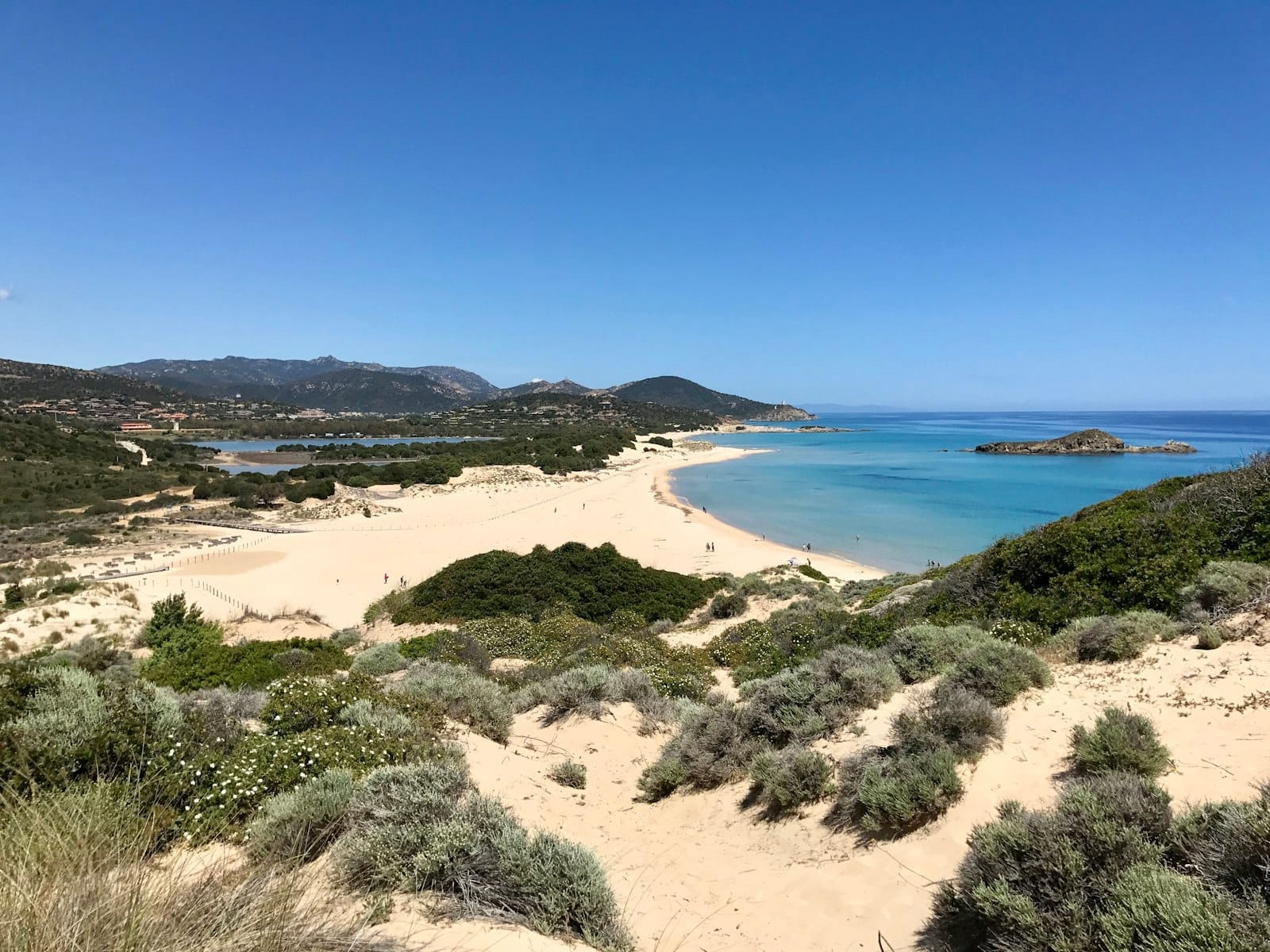 Weitläufiger Blick auf einen unberührten Sandstrand mit türkisfarbenem Wasser, sanften Wellen und mediterranen Sträuchern in der Nähe von Cagliari, Sardinien unter einem klaren blauen Himmel.