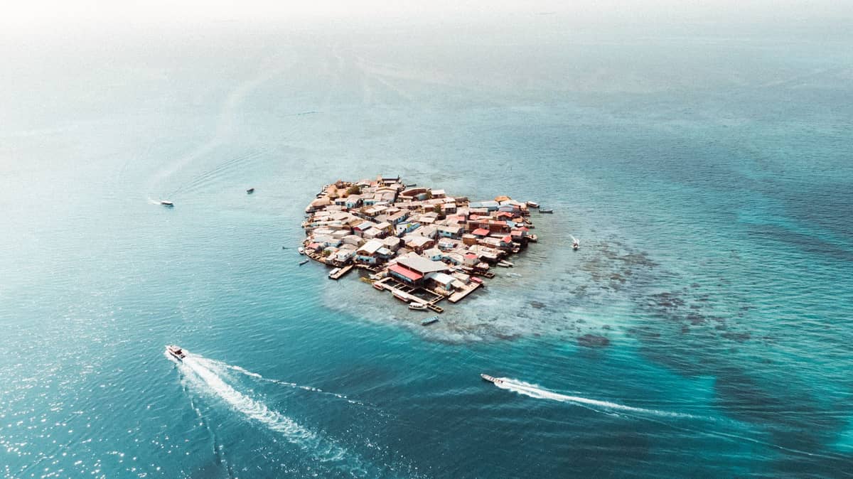 Aerial view of a small island densely packed with colorful buildings, surrounded by turquoise water and boats approaching.