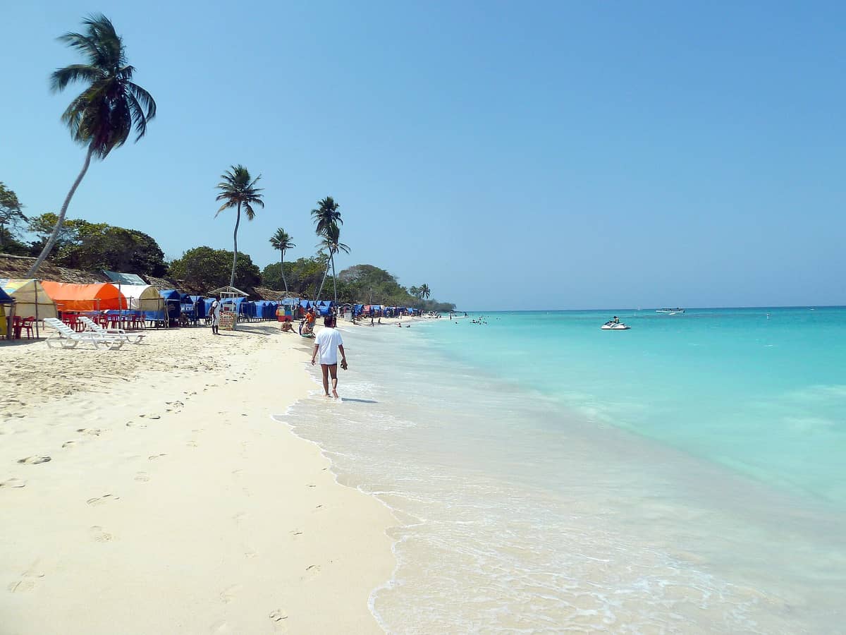 Wide view of Playa Blanca near Cartagena with palm trees, colorful beach tents, tourists, blue sky, and turquoise Caribbean water.