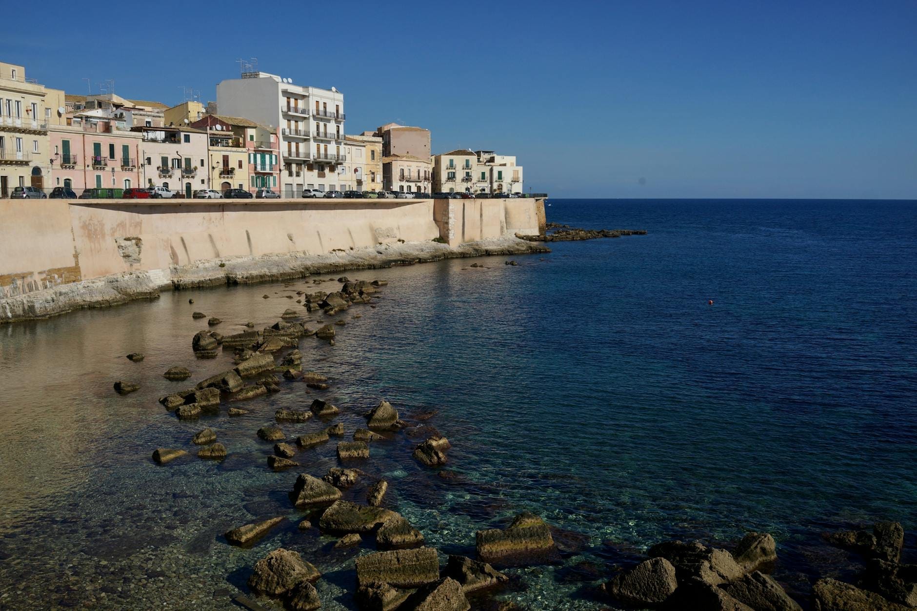 Strandpromenade mit historischen Gebäuden und Stadtmauern am klaren blauen Wasser in Ortigia, Siracusa, unter einem strahlend blauen Himmel.