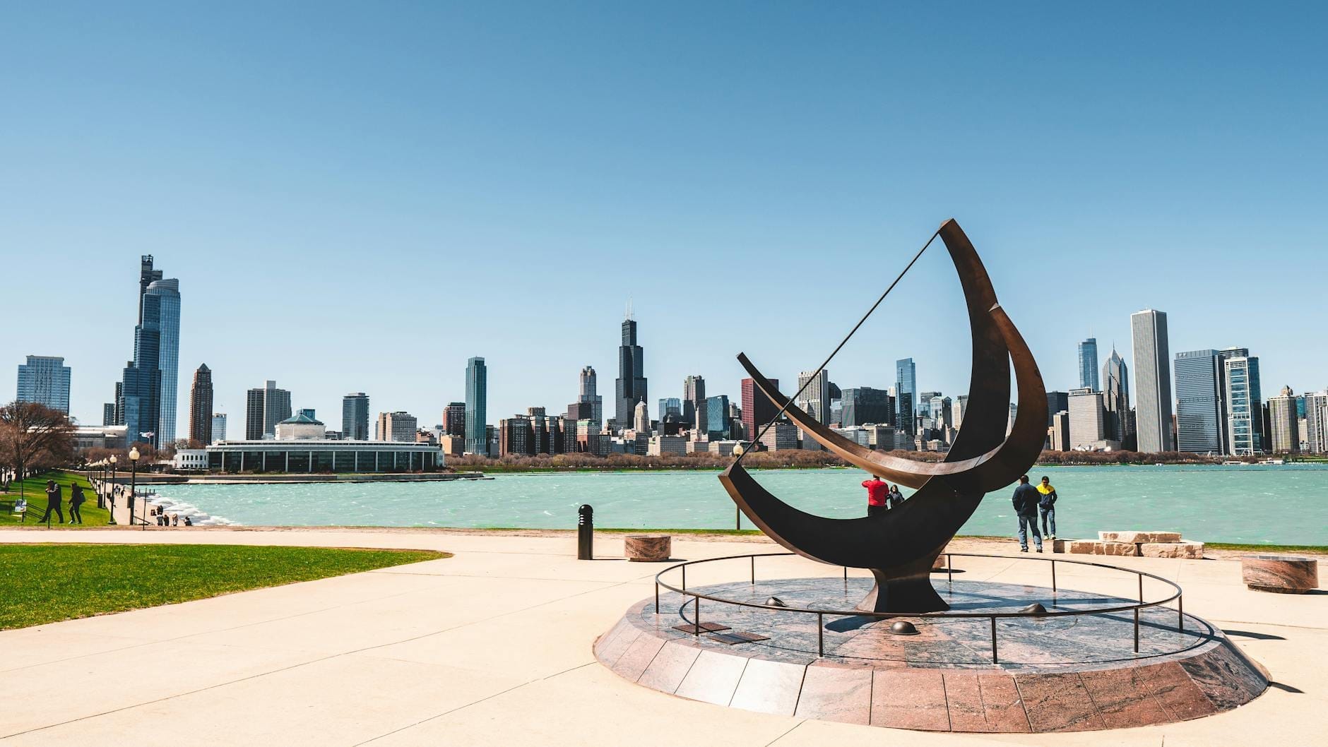 Horizonte de Chicago visto de um parque à beira do lago em um dia ensolarado, com uma grande escultura moderna em primeiro plano e pessoas caminhando pela orla.