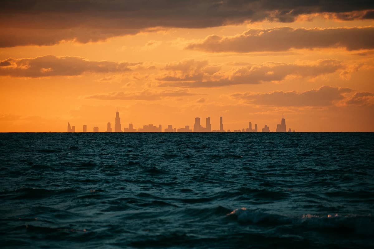 Wellen des Lake Michigan mit der Silhouette der Skyline von Chicago vor einem dramatischen orangefarbenen Abendhimmel.