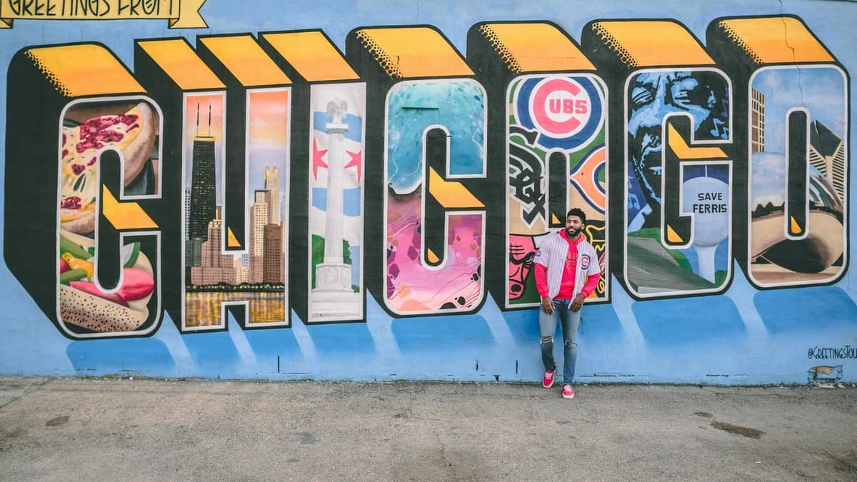 Person, die vor einem großen Wandgemälde mit dem Schriftzug 'CHICAGO', ikonischen Stadtbildern und Symbolen auf einer blauen Wand steht.