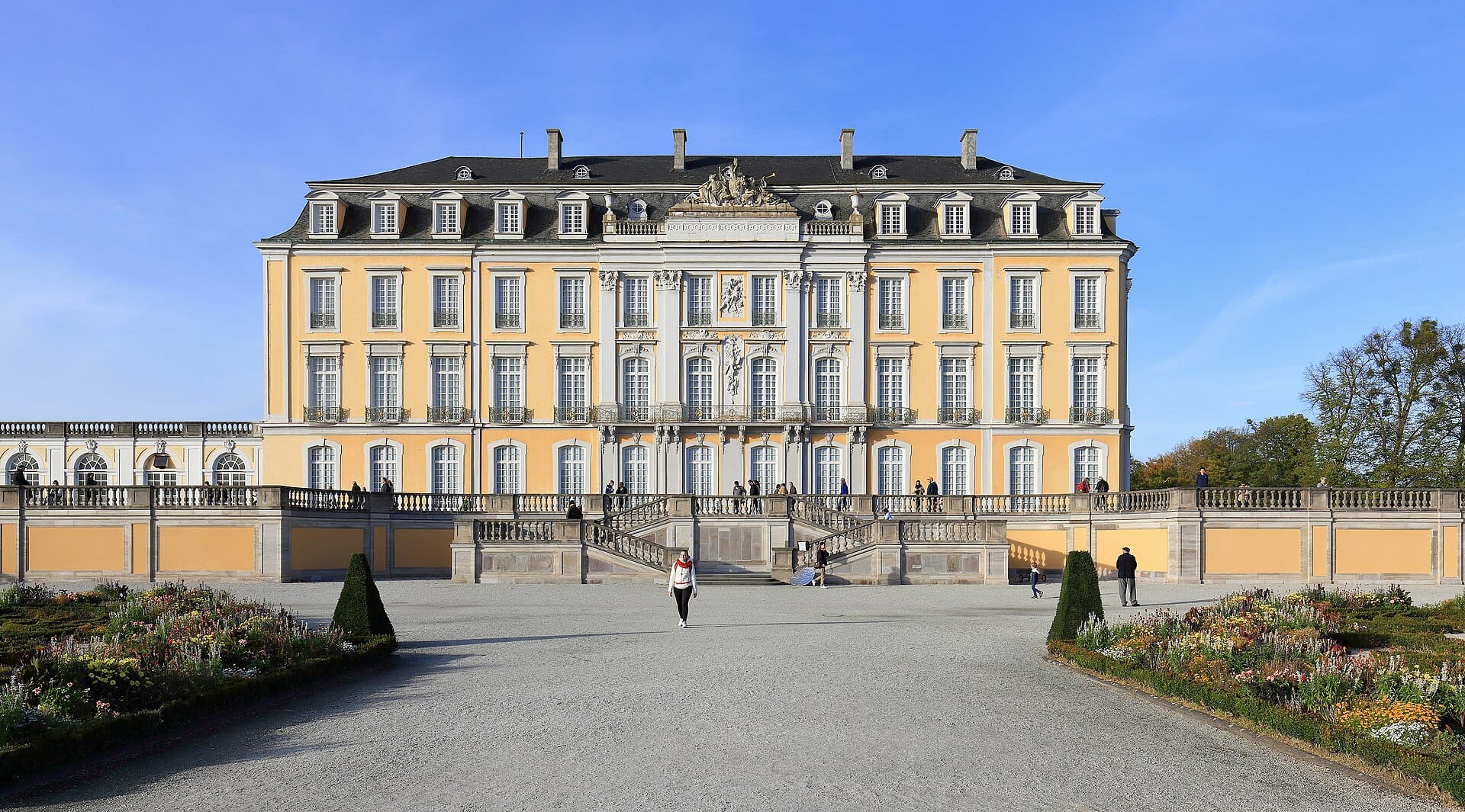 มุมมองด้านหน้าของ Schloss Augustusburg ในเมือง Brühl แสดงให้เห็นด้านหน้าอาคารสไตล์บาโรกสีเหลือง รายละเอียดประดับตกแต่ง แปลงดอกไม้ในสวน และผู้คนที่สำรวจชมในวันที่แดดสดใส