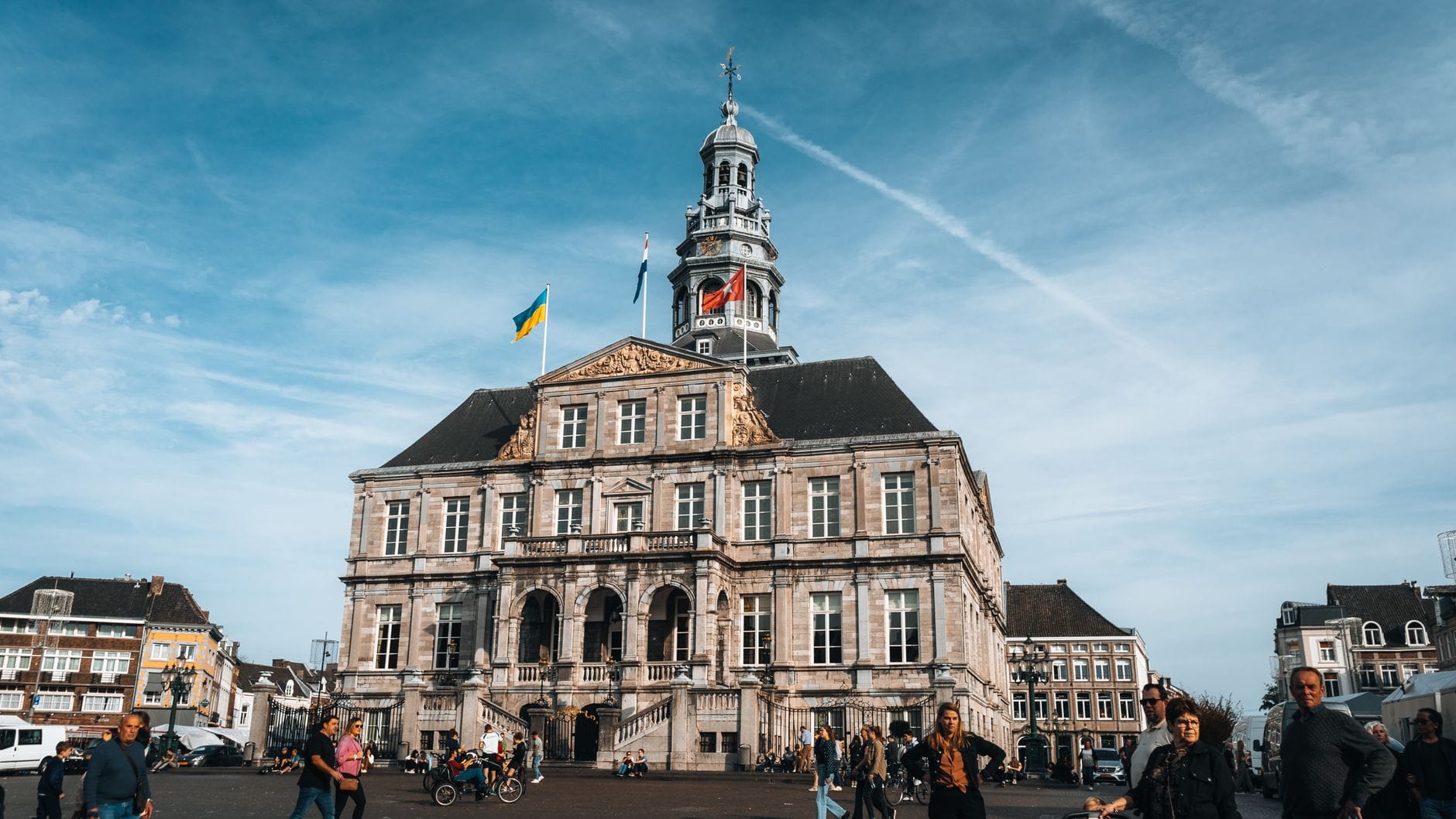 วิวทิวทัศน์ของศาลาว่าการเมืองประวัติศาสตร์ของ Maastricht พร้อมธงชาติ ผู้คนเดินในจัตุรัส และสถาปัตยกรรมดัตช์คลาสสิกใต้ท้องฟ้าสีคราม