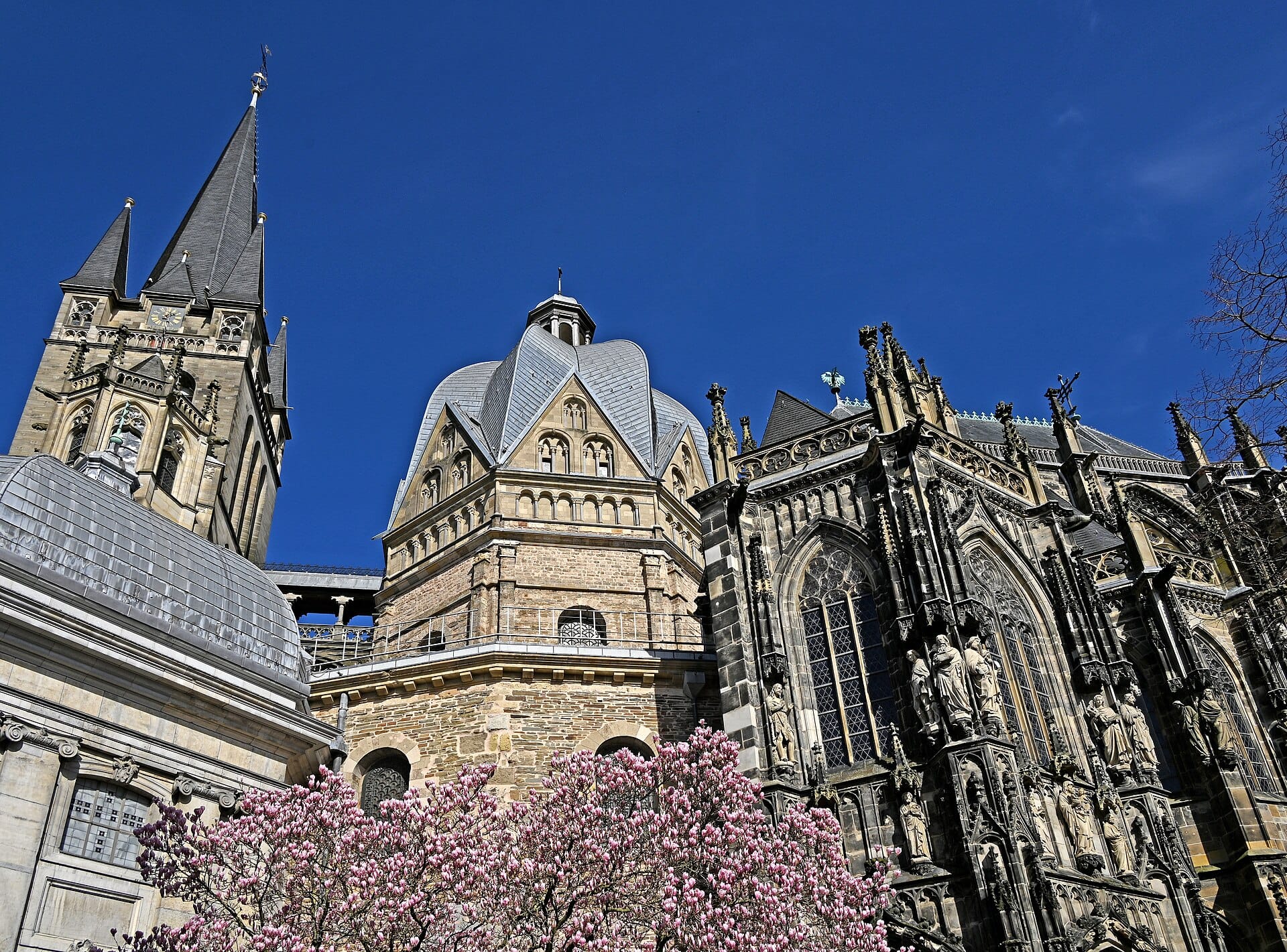 มุมมองกว้างของมหาวิหาร Aachen พร้อมรายละเอียดแบบโกธิกอันวิจิตรและต้นไม้ดอกสีชมพูบานสะพรั่งบนท้องฟ้าสีฟ้าสดใส