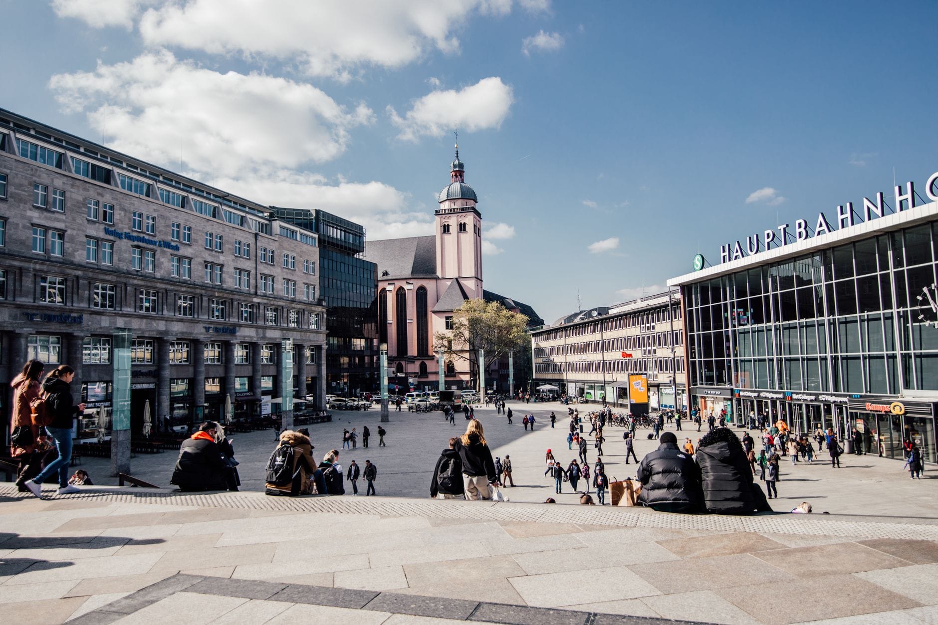 ลานกว้างด้านหน้า Köln Hauptbahnhof มีผู้คนชุมนุมและอาคารในเมืองใต้ท้องฟ้าสีฟ้า