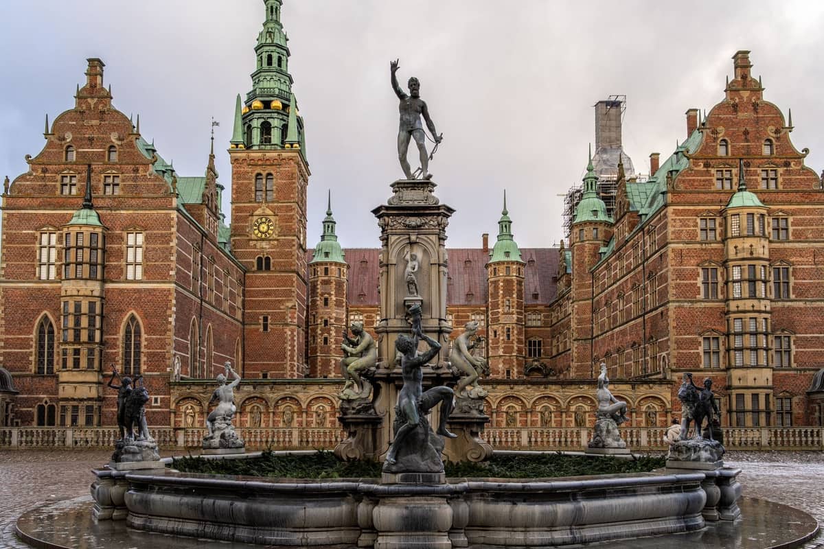 Schlosshof eines großen Renaissance-Schlosses mit verzierten Brunnen und Statuen, roten Backsteintürmen und grünen Kupfertürmen unter bewölktem Himmel.