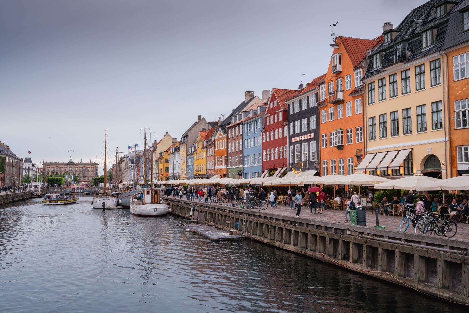 Cena animada de canal em Copenhague com edifícios históricos coloridos, barcos atracados e pessoas passeando à beira-d'água sob um céu nublado.