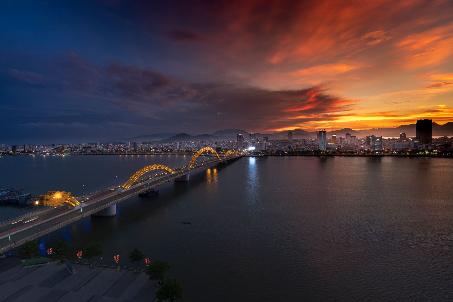 เส้นขอบฟ้าของ Da Nang ยามพระอาทิตย์ตกดิน พร้อม Dragon Bridge ข้ามแม่น้ำและภูเขาเป็นฉากหลัง