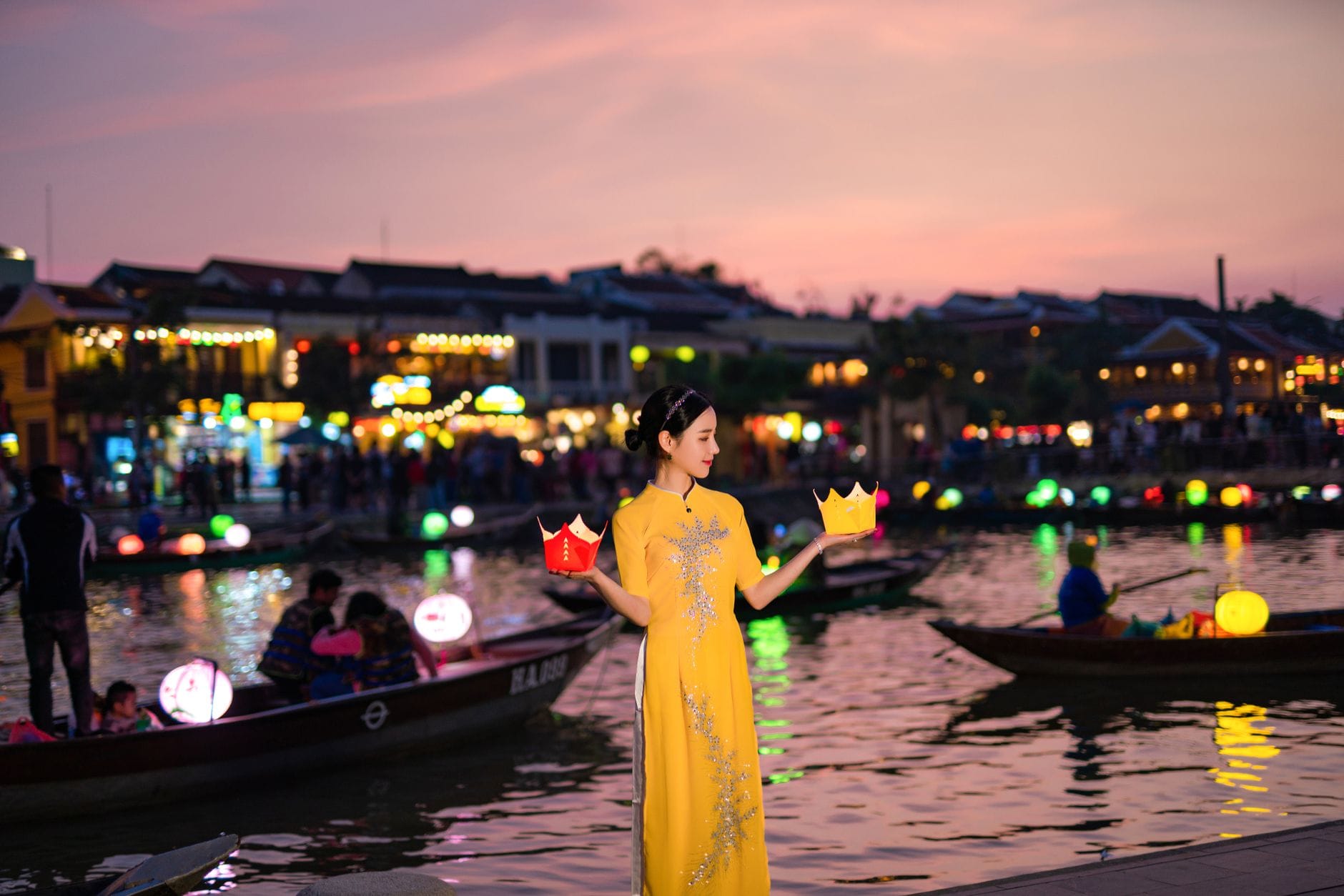หญิงสาวสวมอาวหญิงสีเหลืองถือโคมไฟยามพลบค่ำริมแม่น้ำฮอยอัน พร้อมเรือและเมืองเก่าที่สว่างไสวเป็นฉากหลัง