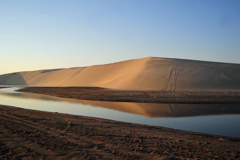 Sanddünen neben dem Inland Sea in Katar, die sich bei Sonnenuntergang unter einem klaren blauen Himmel im Wasser spiegeln.