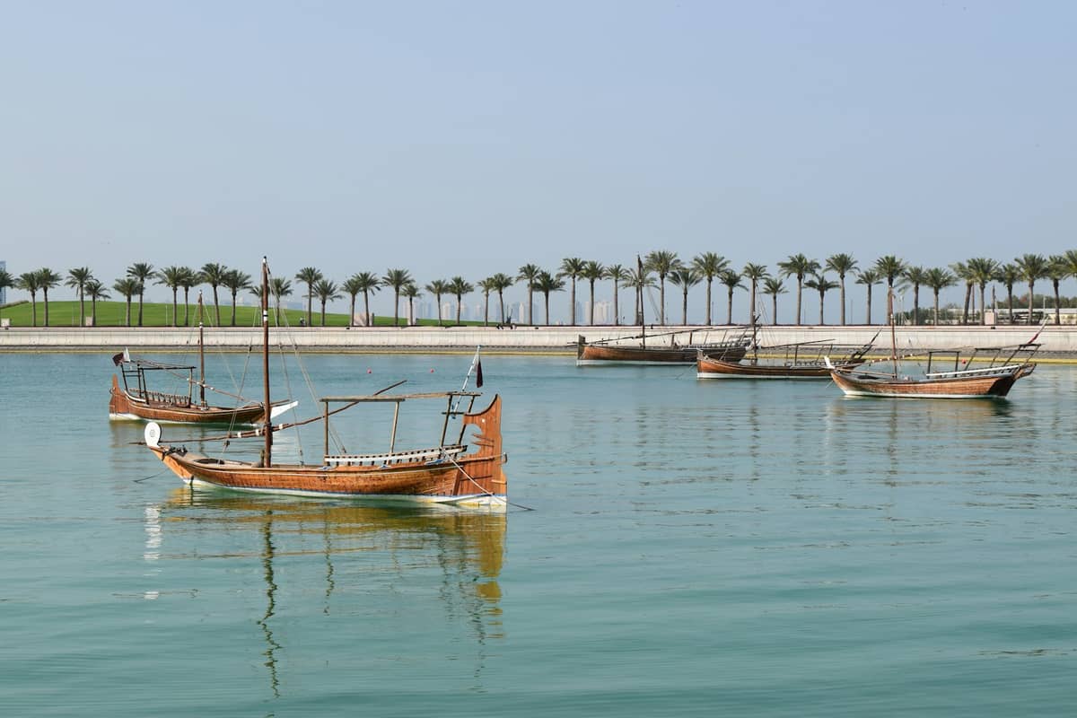 Traditionelle Holzboote auf ruhigem türkisfarbenem Wasser mit Palmen und einem klaren blauen Himmel im Hintergrund nahe der Küste von Doha.