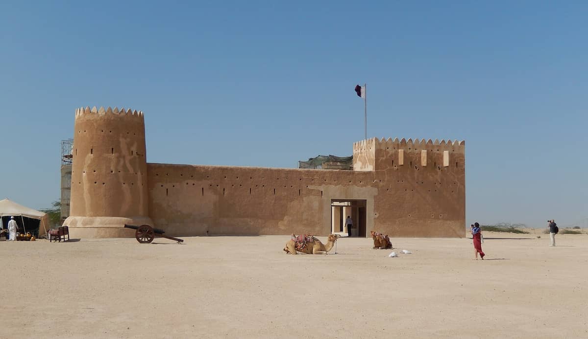 Weitwinkelansicht des Al Zubarah Fort im Norden von Katar, mit Kamelritten und Touristen davor an einem klaren, sonnigen Tag.