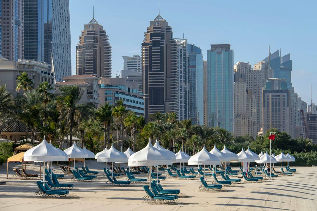 Reihen weißer Sonnenschirme und Liegestühle an einem Sandstrand mit den Wolkenkratzern der Dubai Marina im Hintergrund unter klarem Himmel.