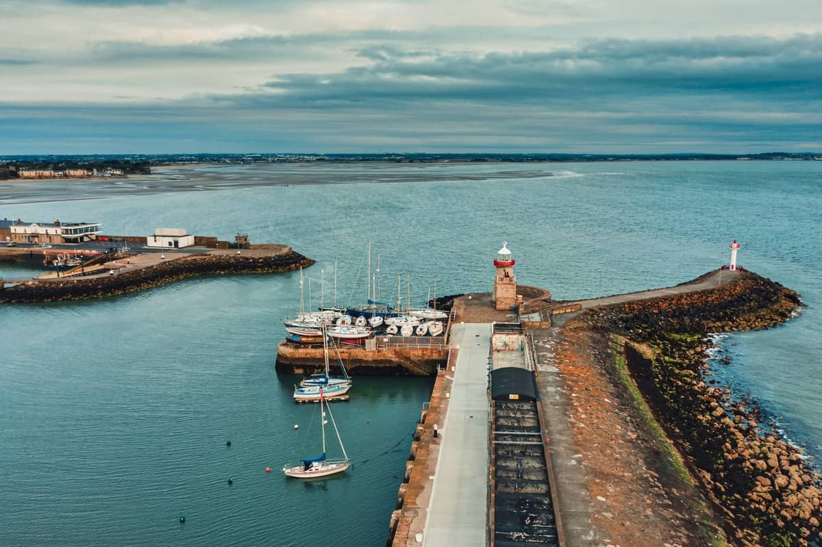 Vista aérea de un puerto con pequeñas embarcaciones, un muelle de piedra y dos faros a lo largo de la costa de Dublín bajo un cielo nublado.