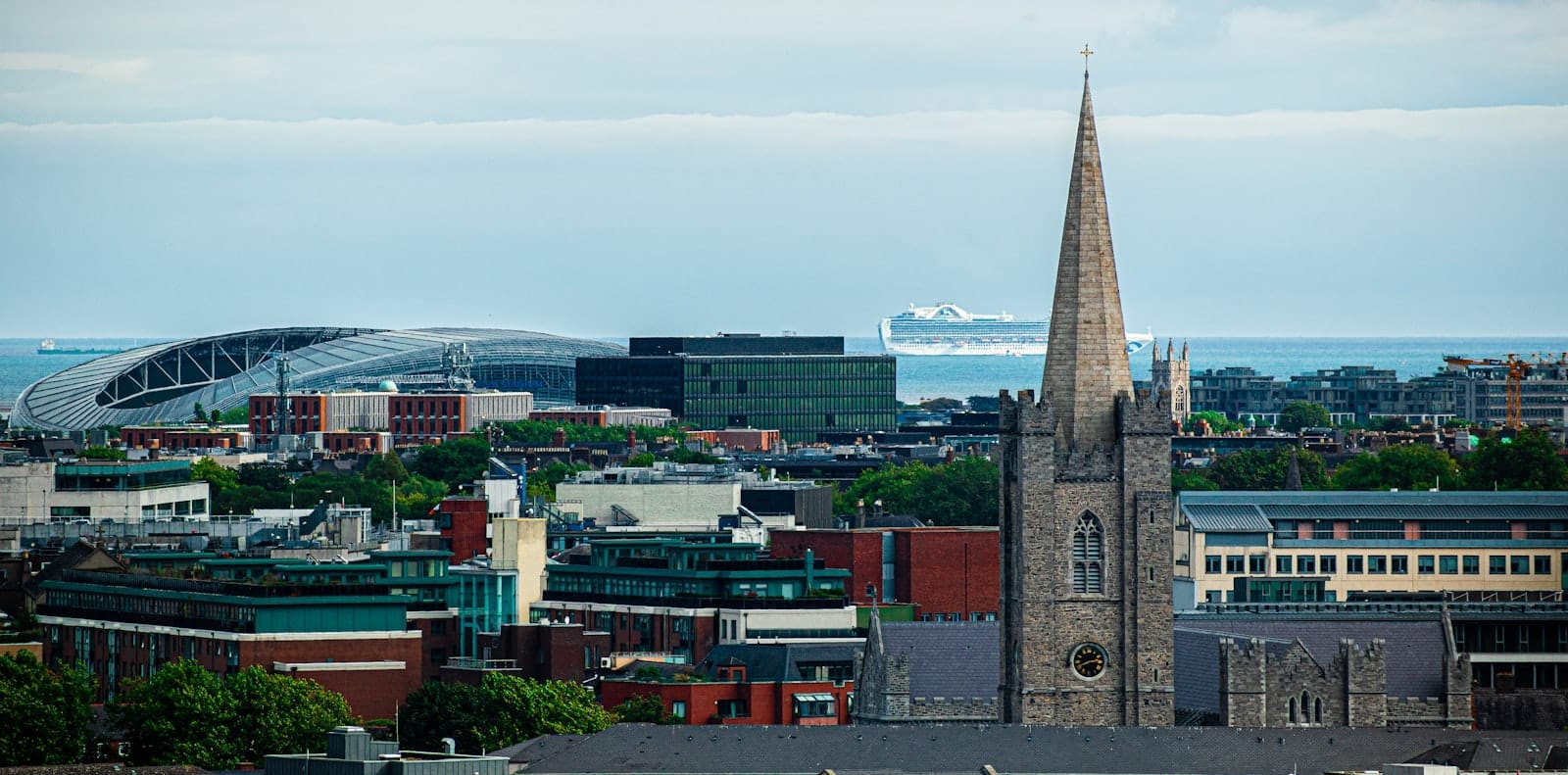 Vista ampla de Dublin mostrando a torre de St Patrick's Cathedral, o Aviva Stadium e uma balsa ao longe no Mar da Irlanda sob um céu azul.