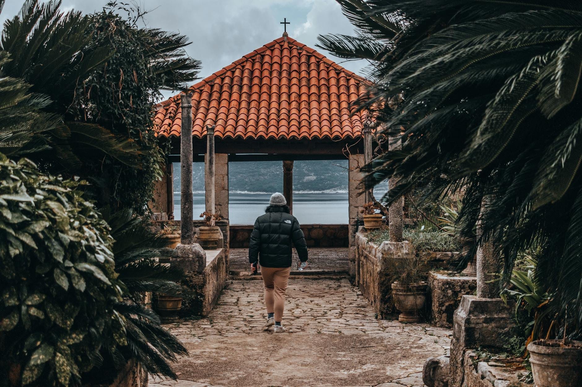 Persona caminando por un exuberante sendero de jardín hacia un pabellón con vistas al mar, rodeado de plantas mediterráneas cerca de Dubrovnik.