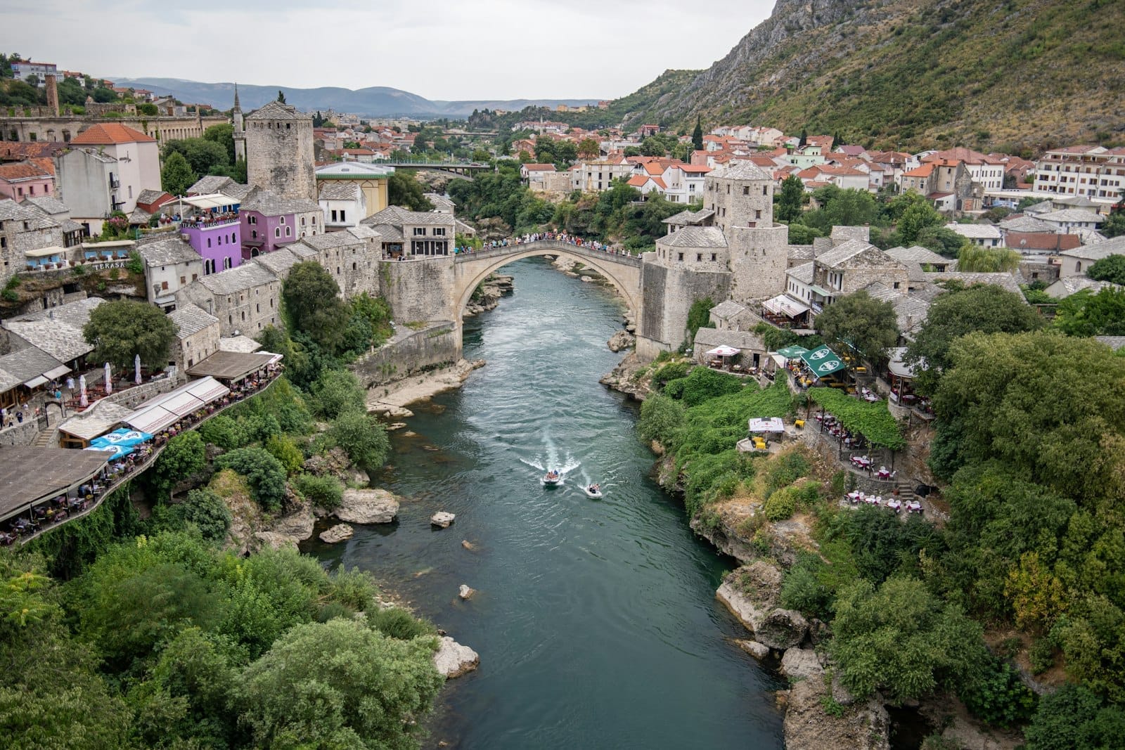 Vista aérea amplia de Mostar con su icónico puente de piedra que cruza el río, rodeado de edificios tradicionales y colinas frondosas.