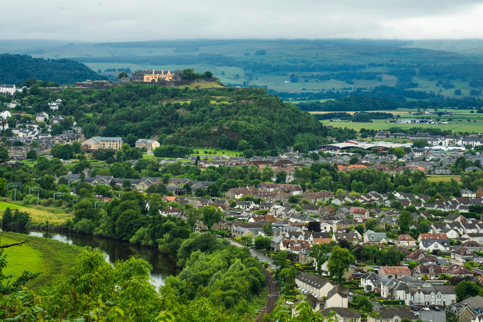Vista panoramica di Stirling con lo Stirling Castle su una collina, circondato da campi verdi, alberi ed edifici della città sotto un cielo nuvoloso.