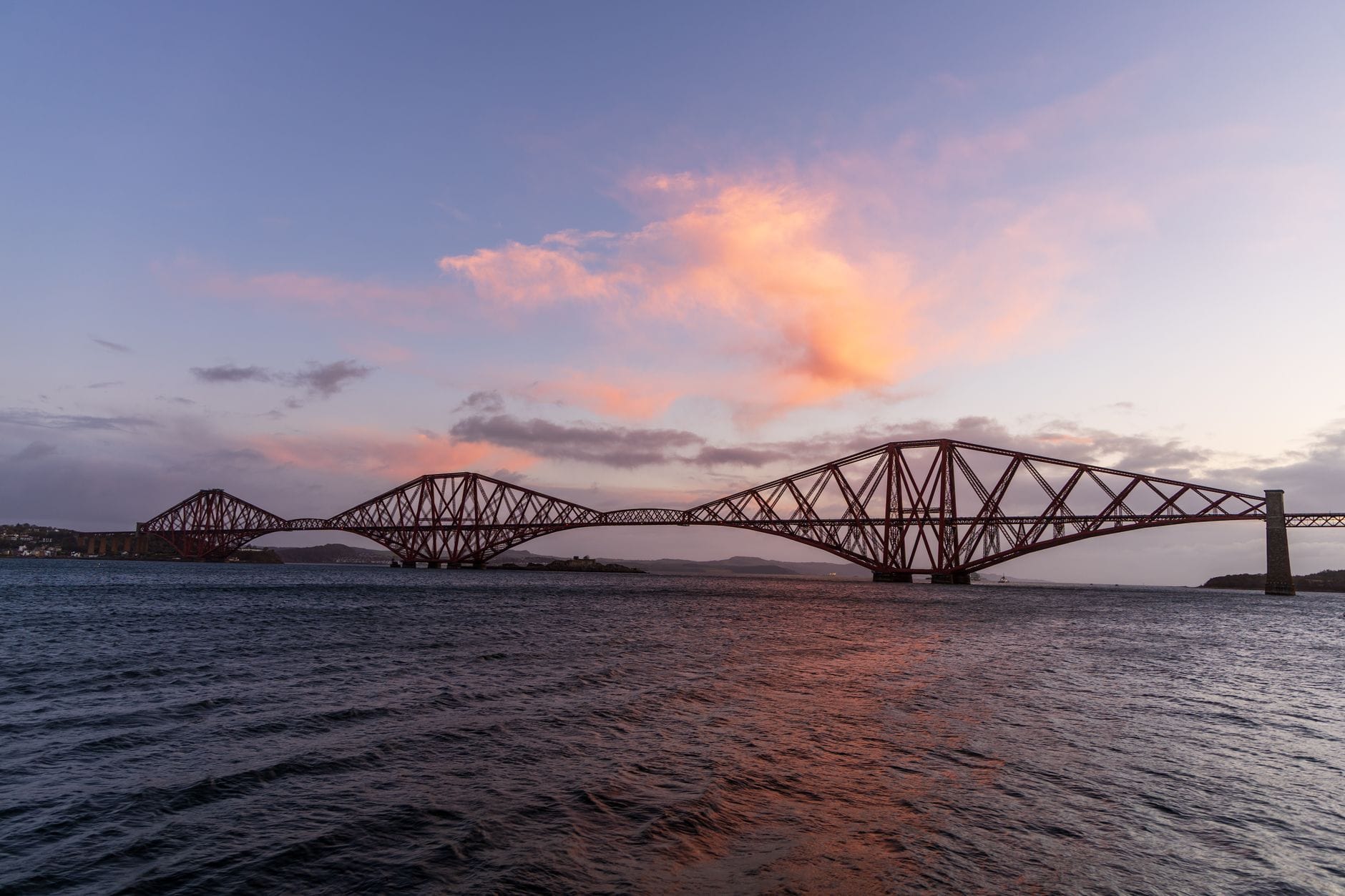 Vista ampia dell'iconico Forth Bridge rosso che si estende sull'acqua al tramonto, con nuvole rosa in alto e onde calme in basso.