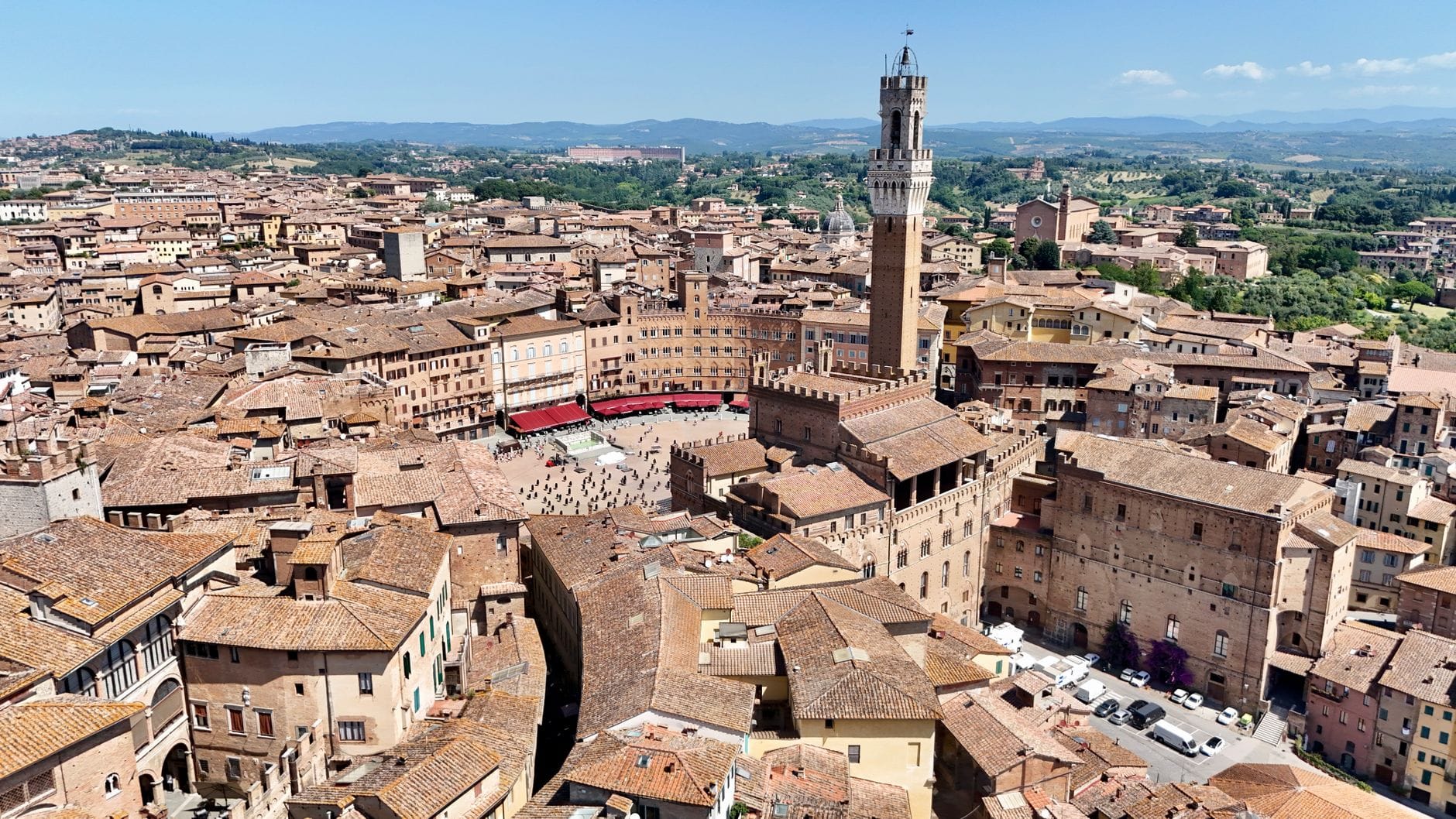ภาพทางอากาศของ Piazza del Campo และ Torre del Mangia ในเมือง Siena พร้อมหลังคาบ้านและชนบท Tuscany เป็นฉากหลังใต้ท้องฟ้าสีฟ้า
