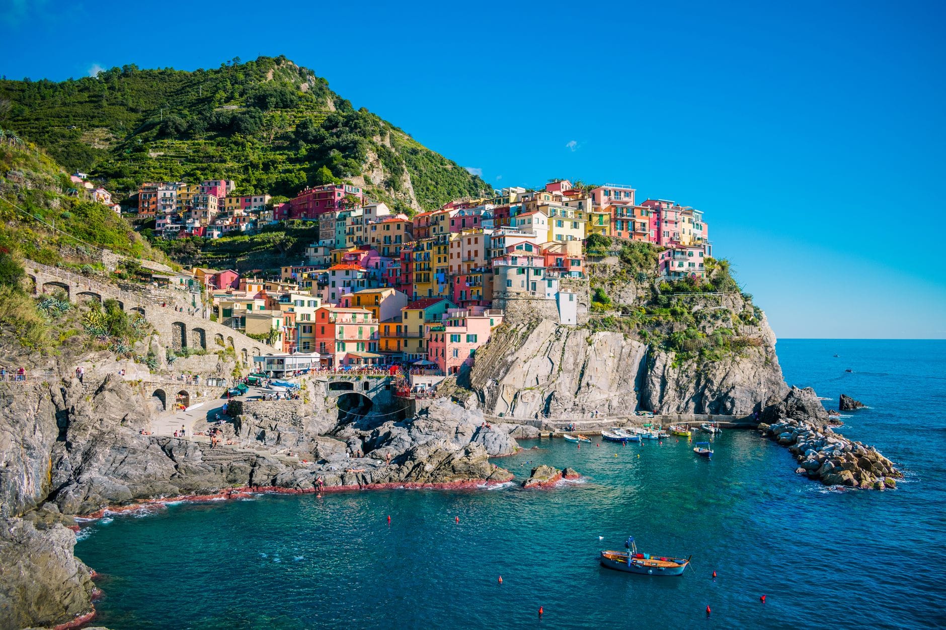 หมู่บ้านริมหน้าผา Manarola ใน Cinque Terre ที่มีสีสันสดใส มองเห็นทะเล Ligurian สีฟ้า พร้อมเรือในน้ำและเนินเขาสีเขียวด้านหลัง