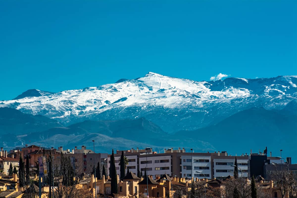 ยอดเขา Sierra Nevada ที่ปกคลุมด้วยหิมะตั้งตระหง่านอยู่เบื้องหลังอาคารในเมือง Granada ภายใต้ท้องฟ้าสีฟ้าใสๆ