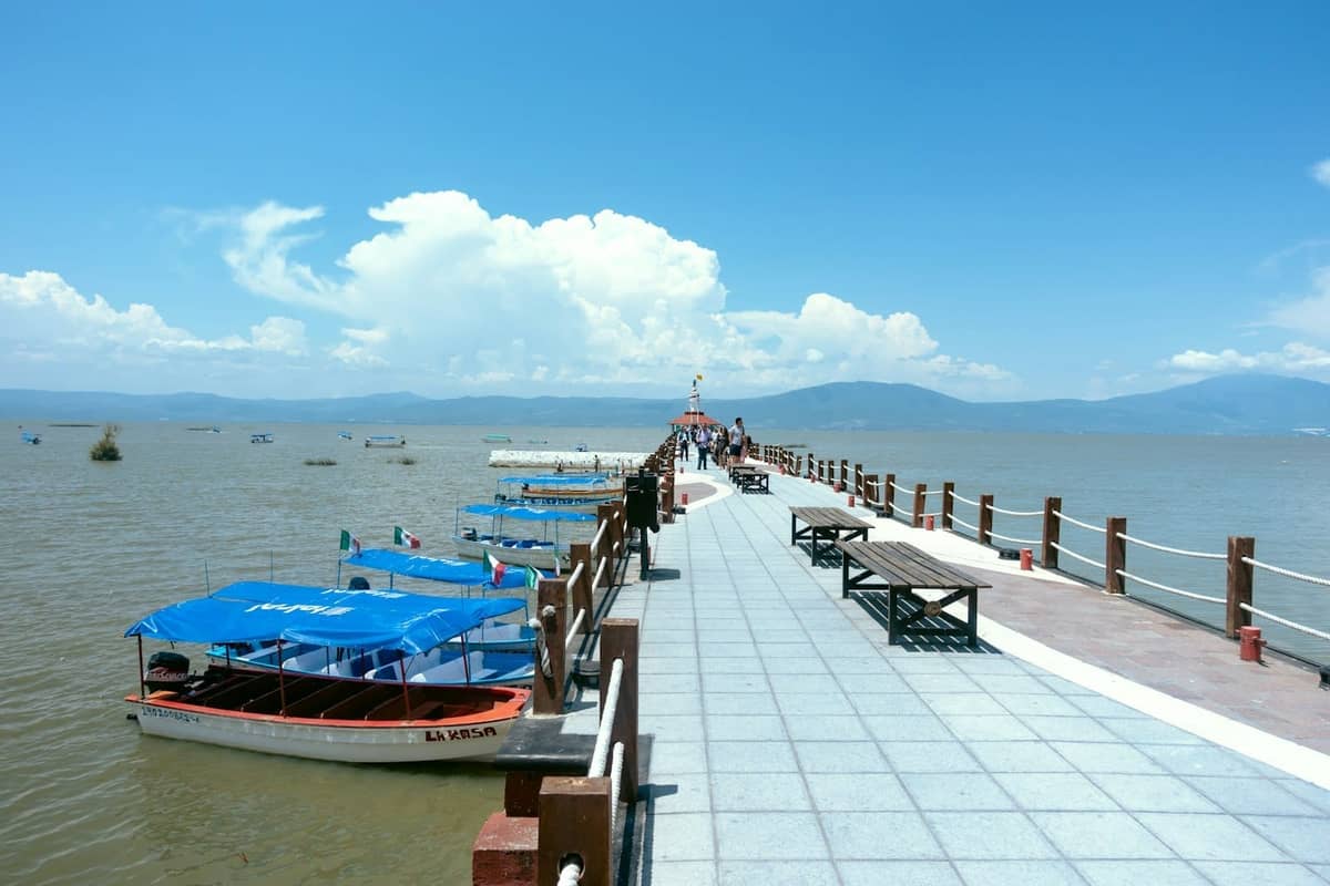 ท่าเทียบเรือริมทะเลสาบที่สดใสทอดยาวเหนือทะเลสาบชาปาลา พร้อมเรือหลังคาสีน้ำเงินจอดเทียบท่า มีภูเขาและเมฆอยู่เบื้องหลัง
