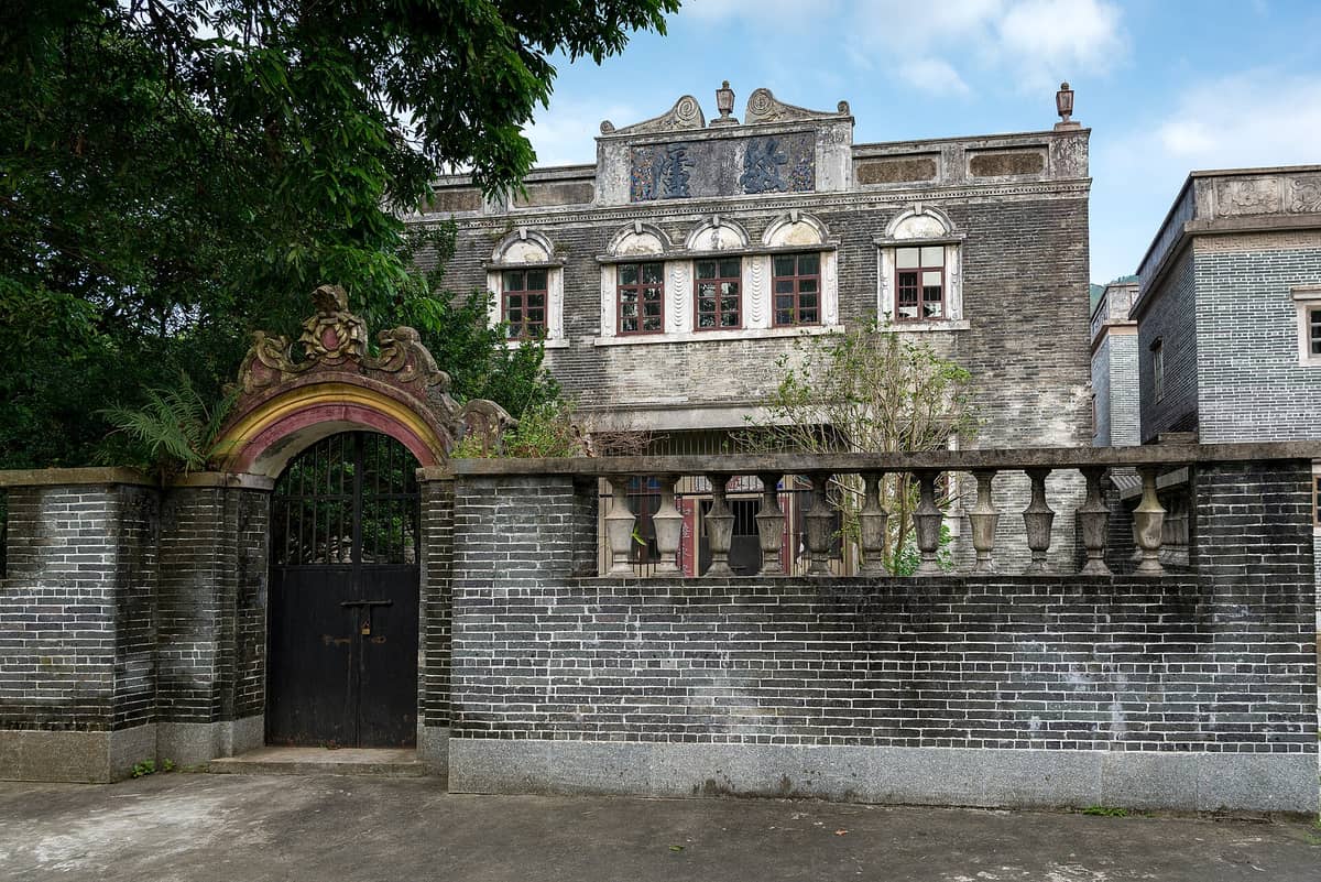 Historic grey brick Kaiping diaolou building with decorative archway and gated entrance, surrounded by stone wall and foliage.