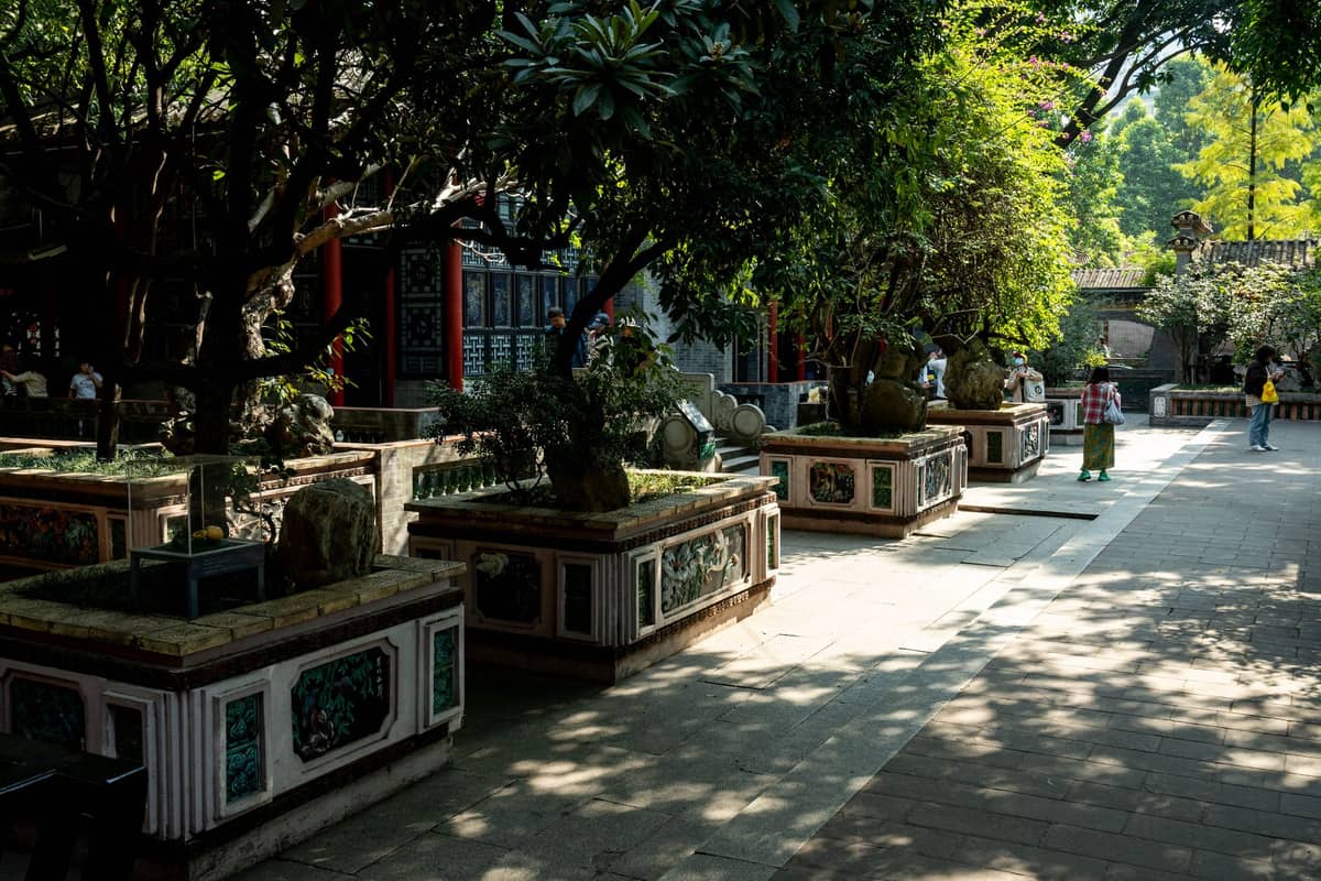 Sunlit courtyard with decorative stone arrangements and traditional architecture, people walking under lush green trees, likely in a historic or cultural site.