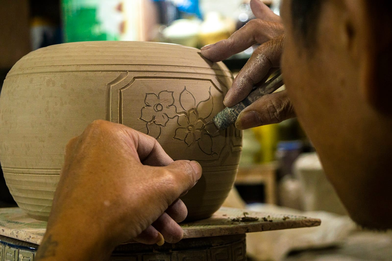 Close-up of artisan hands carving a flower design on a clay pot in a traditional ceramic workshop.