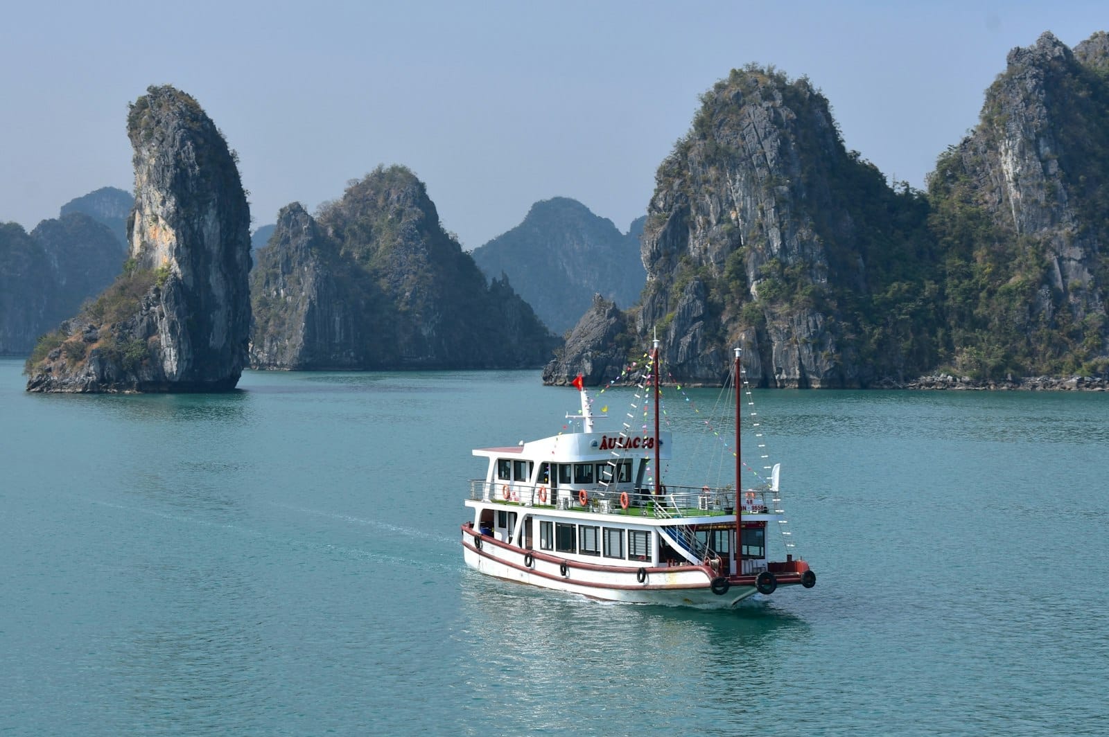 Ha Long Bay with a white cruise boat sailing among limestone karsts on calm blue water under a clear sky.