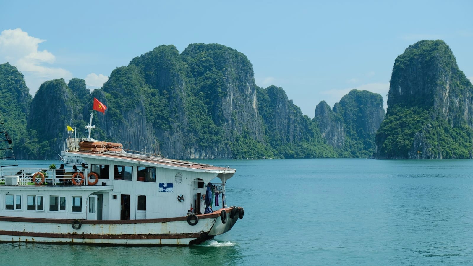 Tour boat cruises across emerald waters with dramatic limestone karsts of Ha Long Bay in the background under a bright, clear sky.